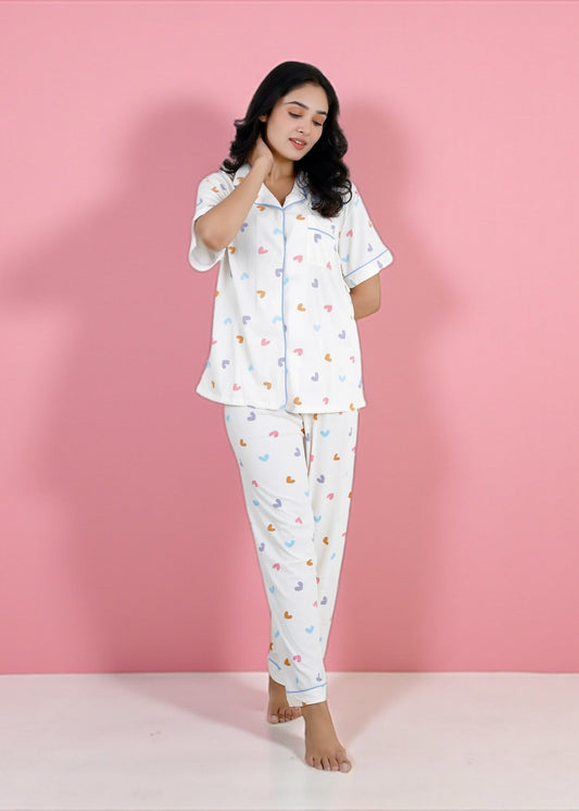 Woman wearing white cotton pyjama set with pastel rainbow hearts and sky-blue piping, full-length pants and short-sleeve shirt, by WakeUp Chicq.