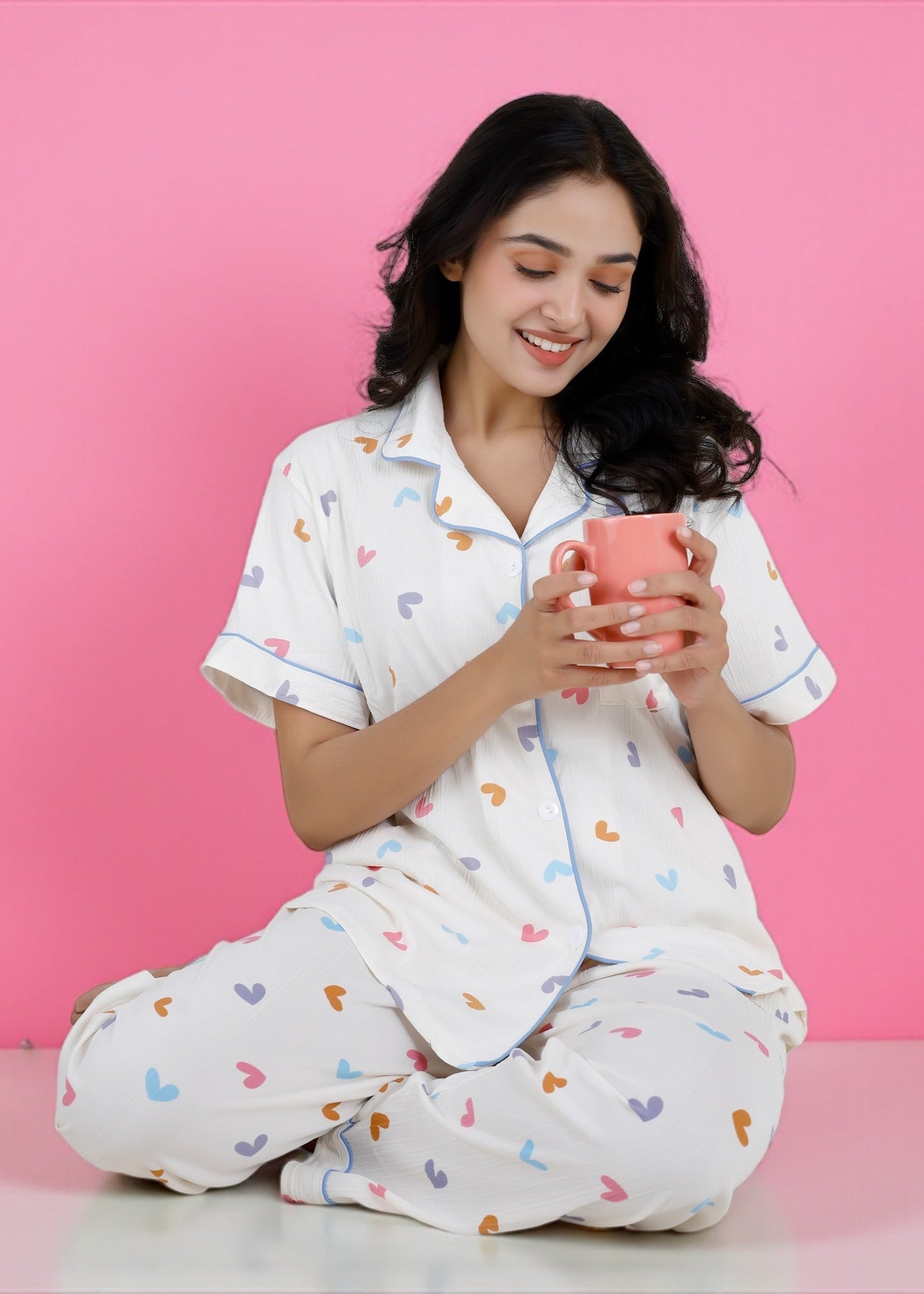 Woman wearing white cotton pyjama set with pastel rainbow hearts and sky-blue piping, full-length pants and short-sleeve shirt, by WakeUp Chicq.
