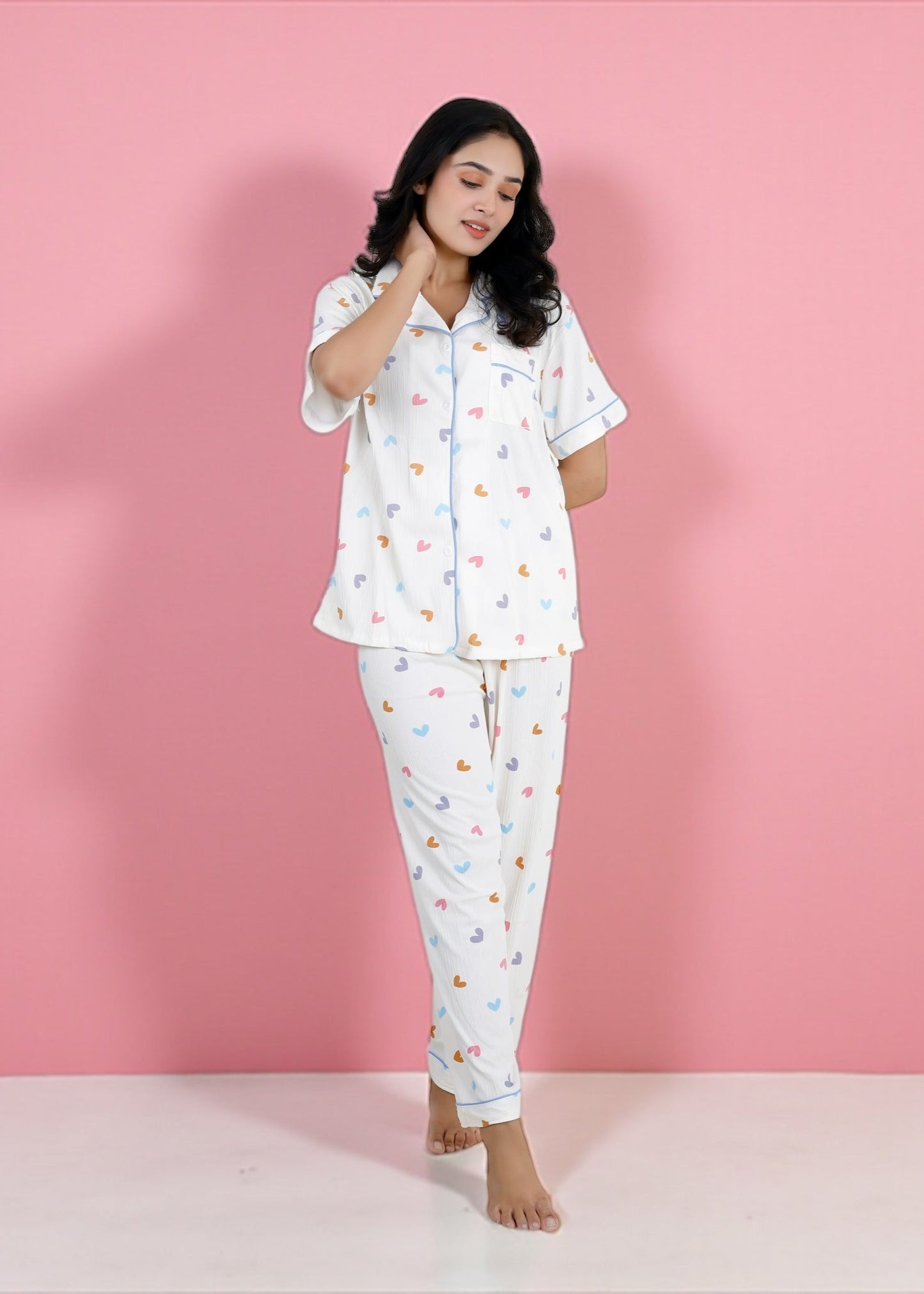 Woman wearing white cotton pyjama set with pastel rainbow hearts and sky-blue piping, full-length pants and short-sleeve shirt, by WakeUp Chicq.