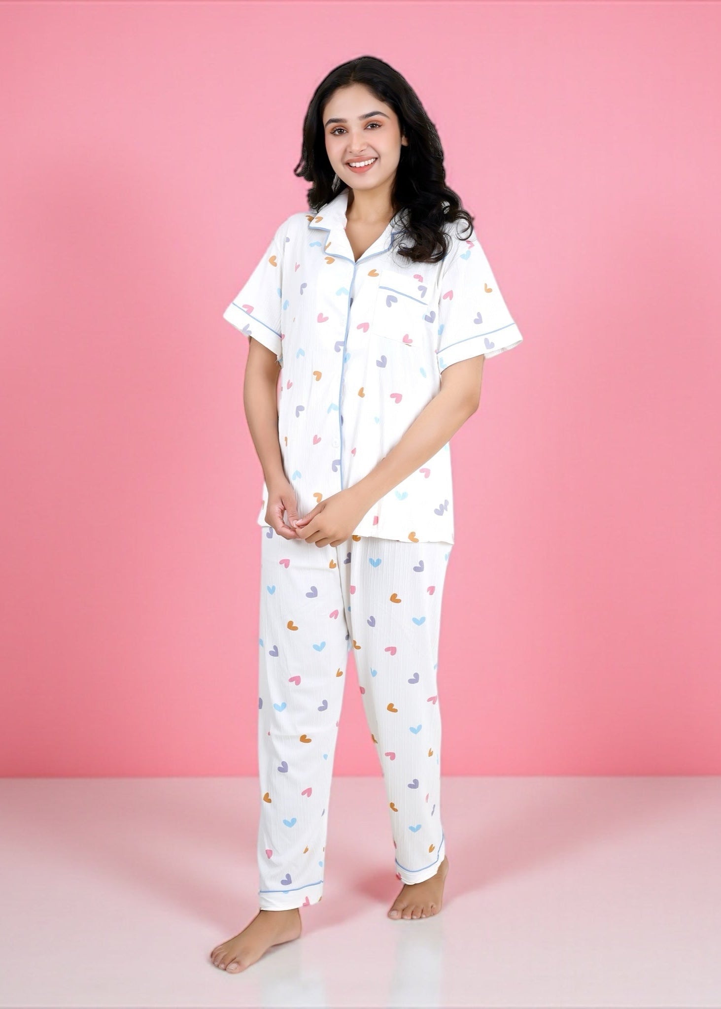 Woman wearing white cotton pyjama set with pastel rainbow hearts and sky-blue piping, full-length pants and short-sleeve shirt, by WakeUp Chicq.